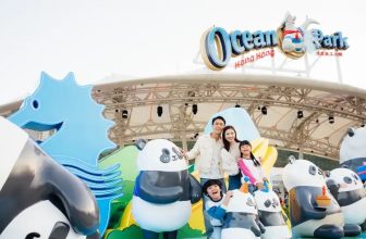 Trip.com 香港海洋公園官方宣傳圖 — 海洋公園吉祥物與遊客在樂園入口合照，背景為高峰樂園及海洋館遊樂設施與彩色招牌，用於 Trip.com 哈囉喂全園祭優惠（Trip.com 官方圖片）