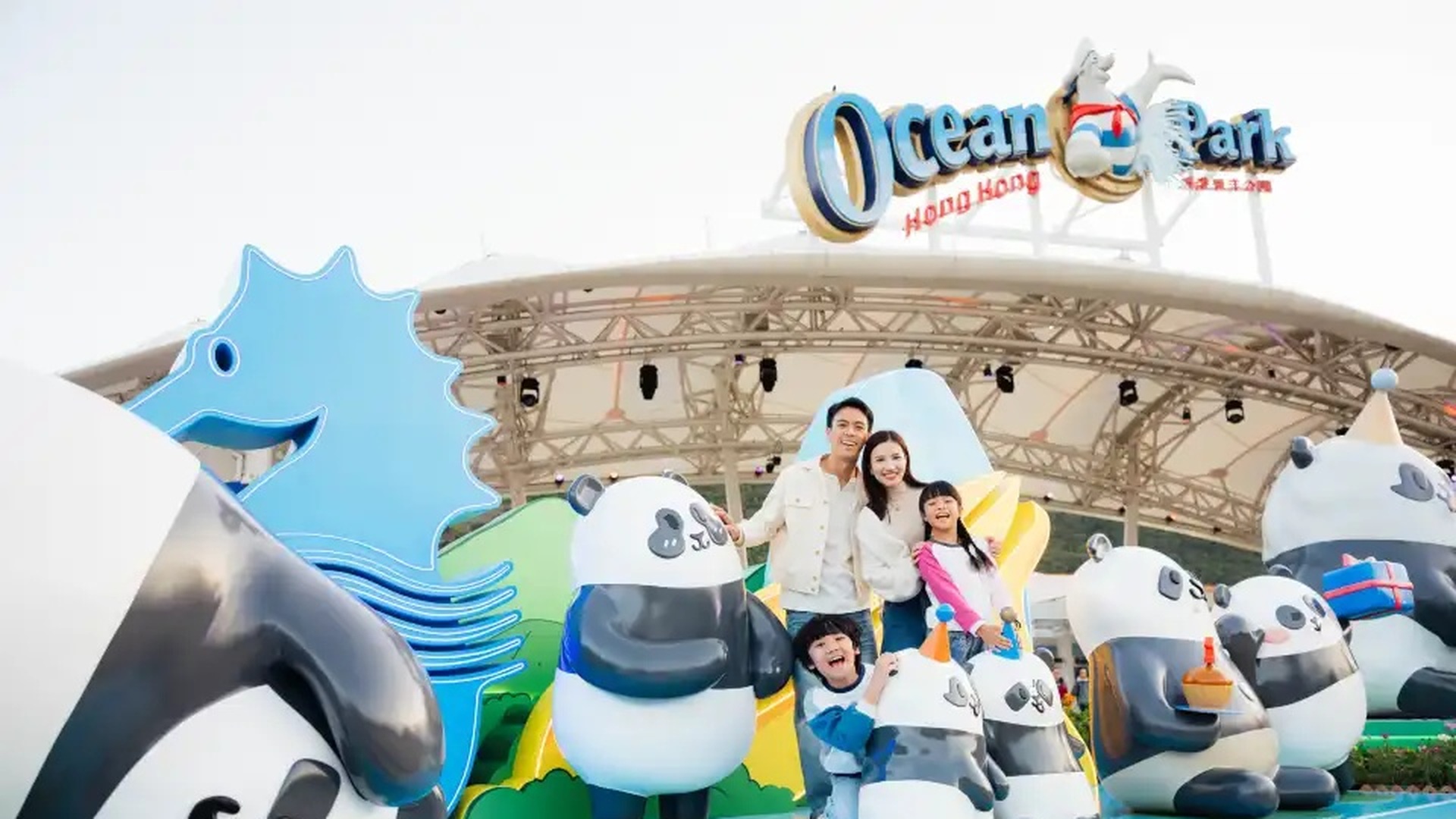 Trip.com 香港海洋公園官方宣傳圖 — 海洋公園吉祥物與遊客在樂園入口合照，背景為高峰樂園及海洋館遊樂設施與彩色招牌，用於 Trip.com 哈囉喂全園祭優惠（Trip.com 官方圖片）
